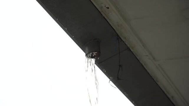 Water drips slowly from a leaking gutter pipe outside on a roof.