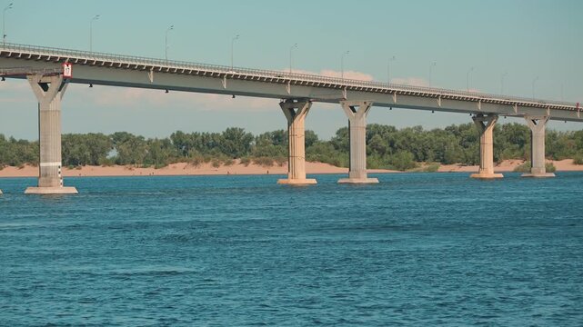 A serene landscape view of a long concrete bridge spanning a wide teal river. Cars cross above while the far bank shows a sandy beach and green trees under a pale blue sky. Calm and peaceful mood
