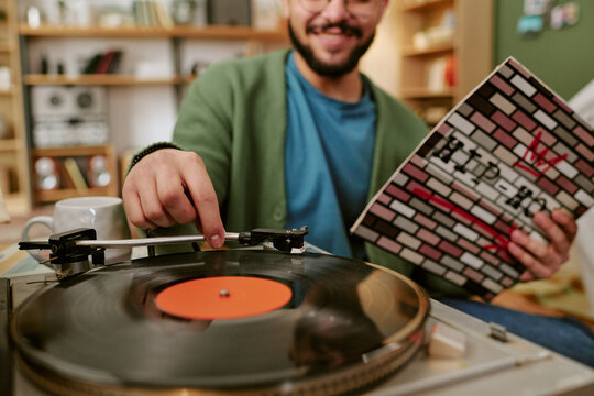 Young adult man playing vinyl record on turntable while holding album cover, enjoying analog music at home