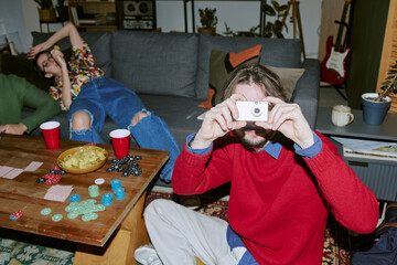 Portrait of Caucasian young adult man holding compact camera and photographing friends during home game night, showing casual social bonding © AnnaStills