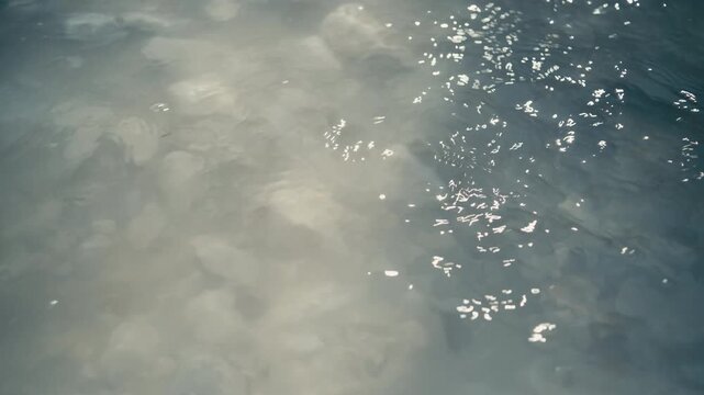 Abstract close-up of sunlight reflections shimmering on a clear water surface with stones beneath. Gentle ripples create dynamic light patterns and a calming natural texture.
