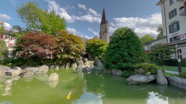 Static shot of a serene Japanese garden with koi fish swimming in a green pond, rocks, lush trees, and a church steeple rising behind under partly cloudy skies in Interlaken, Switzerland.