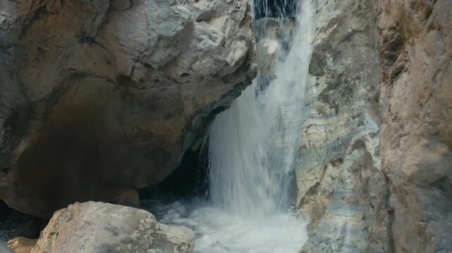 Small waterfall flowing through a rocky canyon with a smooth and colorful stone wall shaped by erosion. Detailed natural textures and mineral patterns create a unique and scenic environment.