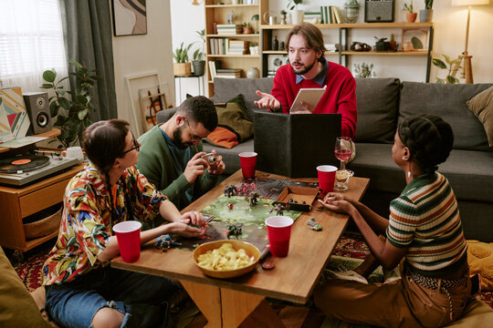 Multiethnic young adults playing tabletop role playing game in living room, discussing strategy and rolling dice