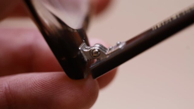 Macro shot of hands holding damaged eyeglasses up close, carefully studying the broken hinge. Fine frame details and wear clearly visible. Real and honest DIY inspection moment.