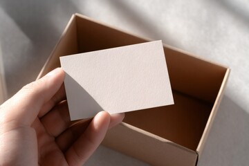 Fototapeta premium Hand holding a blank business card mockup. Empty paper template over a cardboard box for branding identity. Minimalist and professional presentation concept