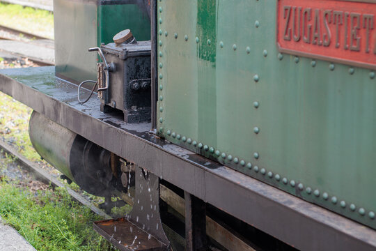 mecanismo de biela y el lubricador goteando de una locomotora de vapor verde vintage, con un fondo de v&iacute;as y hierba, y una placa roja con el texto 'ZUGASTIETA'.