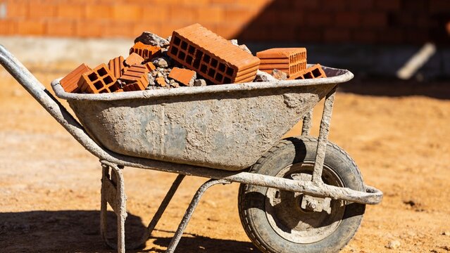 A wheelbarrow filled with red bricks sits on a construction site. The ground is sandy and the background features a brick wall.
