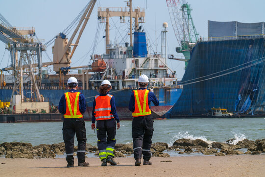 Workers walk along the shore near ships and machinery in a busy port area on a sunny day