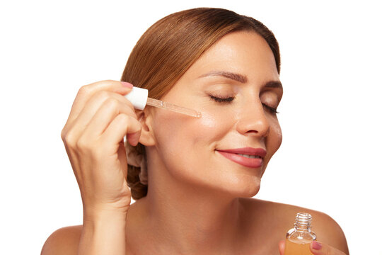 Woman smiling while gently applying skin care serum to her cheek, emphasizing a fresh beauty routine and healthy skin in a well-lit environment.