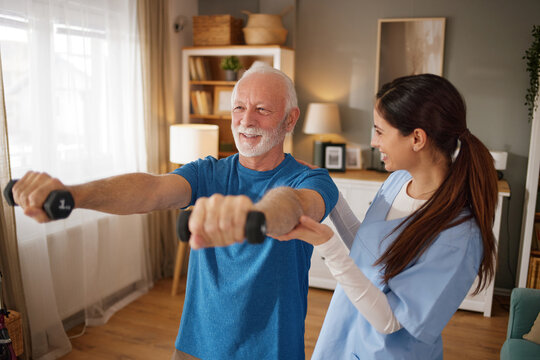 A home health nurse helps an elderly man exercise with dumbbells. They are in a living room setting with natural light coming through the window.