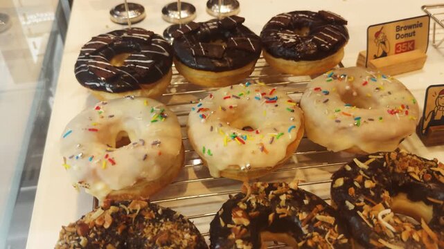 Stacked rows of fresh donuts in a glass display case at a bakery, featuring various toppings like chocolate, glaze, sprinkles, and nuts, attracting customers with a sweet indulgence