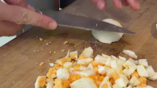 Person chopping hard boiled eggs with a kitchen knife on a wooden cutting board for a healthy protein rich meal, preparing an ingredient for a salad or sandwich