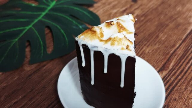 Slice of decadent chocolate layer cake with a rich dark ganache, toasted meringue top, and white icing dripping down, sitting on a plate and wooden surface with a tropical leaf accent