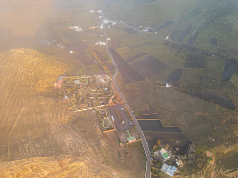 Aerial view of the volcanic landscape with terraced hillsides, a winding road, and traditional architecture under soft golden light in Lanzarote, Canarias, Spain.