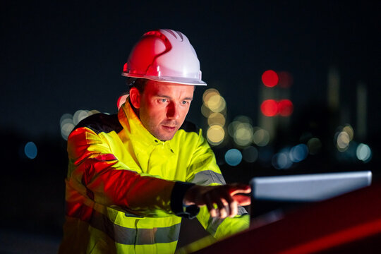 Man in a white hard hat and yellow high-visibility jacket uses a tablet. Serious supervisor monitoring oil refinery operations on a computer late at night with illuminated background.