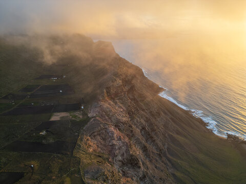 Aerial view of steep volcanic cliffs and agricultural terraces under golden sunset light and rolling mist in Lanzarote, Canarias, Spain.