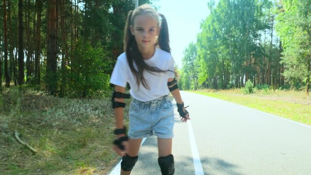 Child girl in casual clothes roller skating on road in forest on sunny summer day.