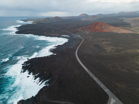 Aerial view of a winding coastal road traversing dark volcanic lava fields and a red cone near crashing waves in Lanzarote, Canarias, Spain.