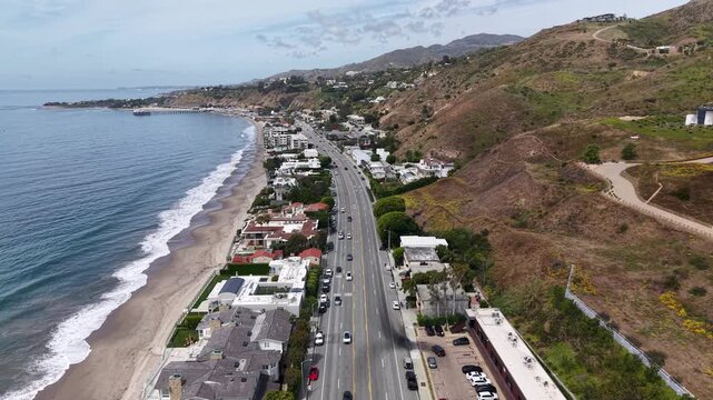 Cinematic 4K 10-bit aerial drone footage captured above the Pacific Coast Highway in Malibu, featuring a smooth reverse dolly movement over vehicles traveling along the scenic coastal highway.