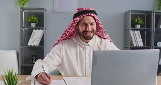 Positive handsome Arabic businessman with beard wearing keffiyeh and thobe online call at laptop, Arab business leader participating in virtual meeting, talking, waving and looking engaged in chat