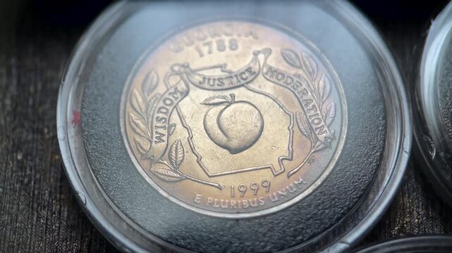Close up video of a 1999 United States quarter dollar coin featuring the Georgia state reverse design with a peach, state outline, and the motto &ldquo;Wisdom, Justice, Moderation.&rdquo;
