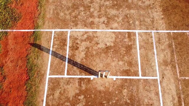 Top-down drone zoom onto cricket stumps and bails on a dirt pitch crease. High-angle overhead shot of sports equipment in bright sunlight.