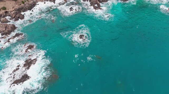 Birds eye view of clear turquoise mediterranean water hitting the rugged cliffs and rocky coastline of Pedra Longa, Sardinia.