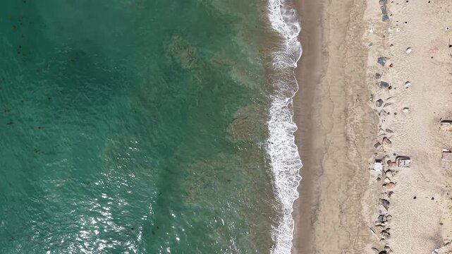 Cinematic 4K 10-bit aerial drone footage captured above Malibu Beach on a sunny day, featuring a bird&rsquo;s-eye view of ocean waves crashing onto the sandy shoreline as the drone slowly dollies forward.