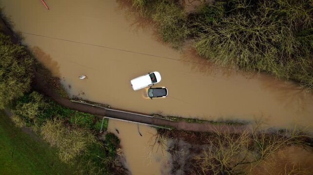 Flash flood disaster leaving cars submerged in muddy water across rural road. Torrential rain creating dangerous high water levels and isolating vehicles near small bridge. Severe weather emergency