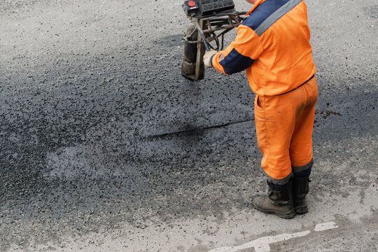 Municipal road worker applies hot bitumen to damaged asphalt seam during pavement repair on city street, close up view.