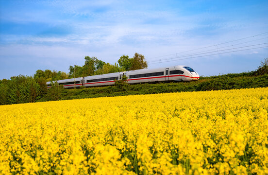 A landscape in the Ruhr region near Bochum-Wattenscheid, featuring yellow fields of rapeseed and a blue sky in warm evening light. A high-speed train from Dortmund to Essen passes along.