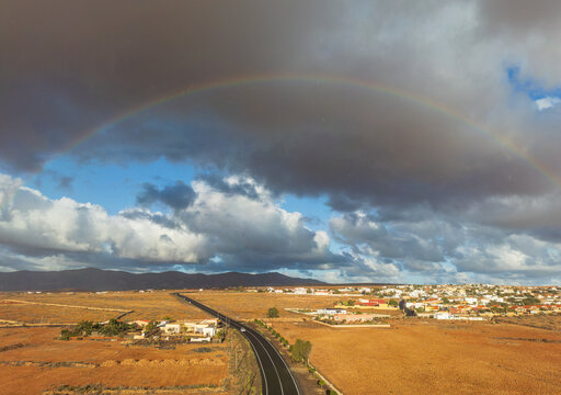 Aerial view of a rainbow arching over a winding road through the volcanic landscape and white houses under a dramatic cloudy sky Lanzarote, Canarias, Spain.