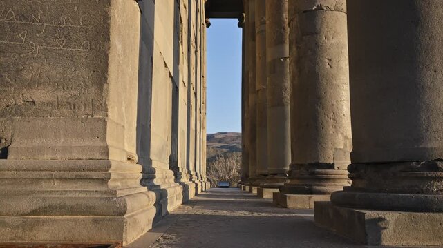 Ancient stone columns line a long corridor. Sunlight glares through gaps between pillars