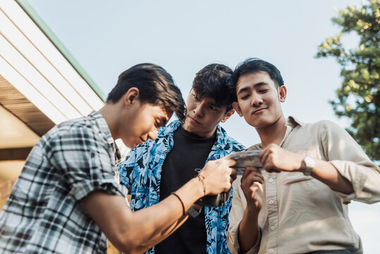 A hesitant young Asian man handing money to two intimidating men during a tense outdoor confrontation. Outdoor shot.