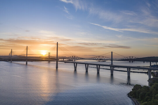 Aerial view of the Forth Bridges including the Queensferry Crossing and the historic red Forth Bridge at sunrise over the Firth of Forth in West Lothian, UK.