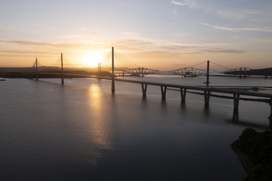 Aerial view of Forth Bridges spanning the Firth of Forth with the sun setting over the calm water and silhouettes of the structures West Lothian, UK.