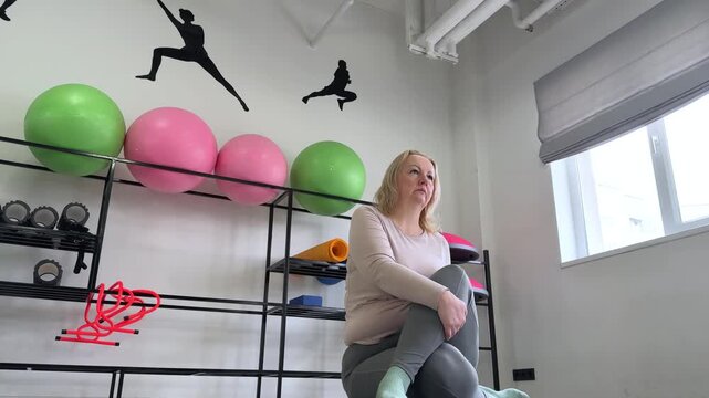 A middle aged woman performs a lying spinal twist on a gym mat, gently rotating her legs to one side while turning her upper body in the opposite direction. The scene is set in a fitness studio.