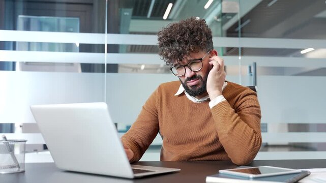 Unhealthy businessman suffers from ear pain sitting at desk at workplace in business office. Male touches his ear with his fingers. Worker has furuncle, shingles, eczema, ear diseases or otitis media