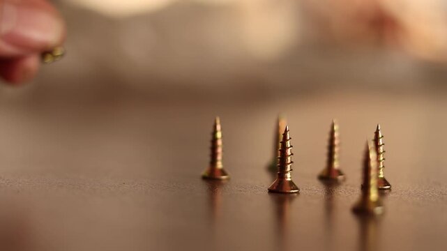 Extreme macro footage of a hand carefully placing a golden wood screw next to a row of standing screws on a polished wooden desk.