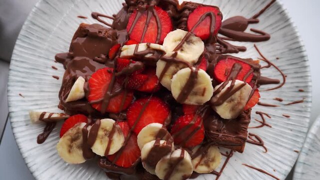 Close-up top down circular view massive, high-calorie waffle topped with strawberries, bananas, and chocolate at an outdoor cafe. A delicious and indulgent big portion sweet treat