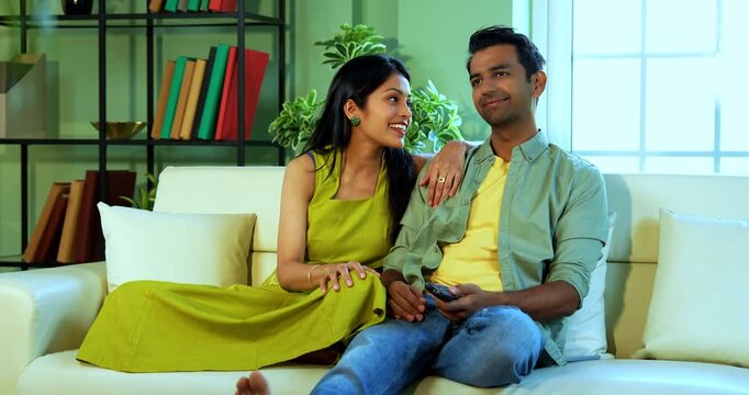 Indian asian young couple watching TV with remote on lazy weekend sitting on sofa in modern Indian home while relaxing and enjoying entertainment together in a comfortable living room setup