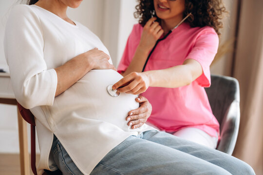 Pregnant woman in white top sitting on chair while female doctor in pink scrubs uses stethoscope to listen to baby's heartbeat in a bright, modern clinic