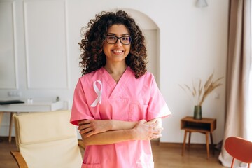 Female healthcare professional wearing pink scrubs with a cancer awareness ribbon stands confidently in a bright, modern office space with plants and furniture visible © standret