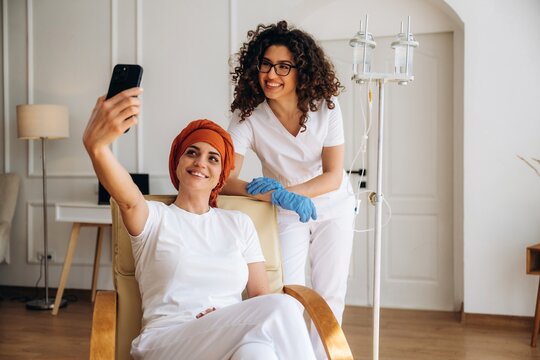Female cancer patient with headscarf taking selfie with smiling nurse in hospital room, IV stand visible, bright interior with wooden flooring and modern decor