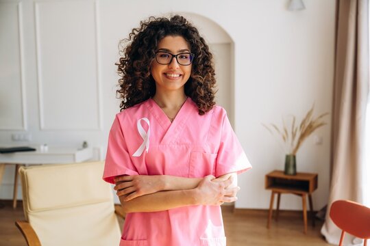 Female healthcare professional wearing pink scrubs with a cancer awareness ribbon stands confidently in a bright, modern office space with plants and furniture visible