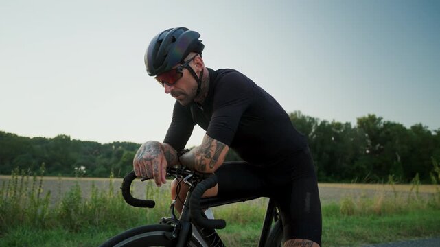 Cyclist Resting Outdoors. Tattooed Male Cyclist Leans On Bike During Sunset In Tranquil Countryside