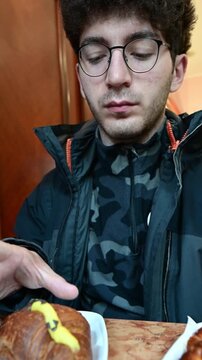 Young man with glasses sitting at a table in a cafe, eating a traditional italian cream croissant for breakfast, enjoying a moment of relaxation during his daily routine in slow motion