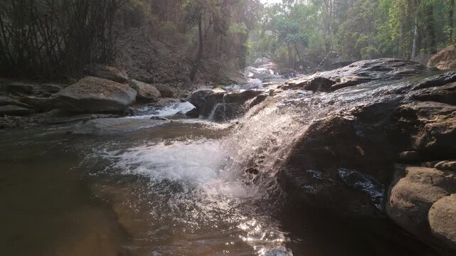 Slow motion of Peaceful stream flowing over rocks in a forest, surrounded by lush greenery and sunlight filtering through the trees.