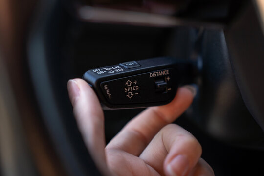 Close-up of hand using cruise control stalk in modern car, showing speed setting and distance adjustment controls on steering column lever.